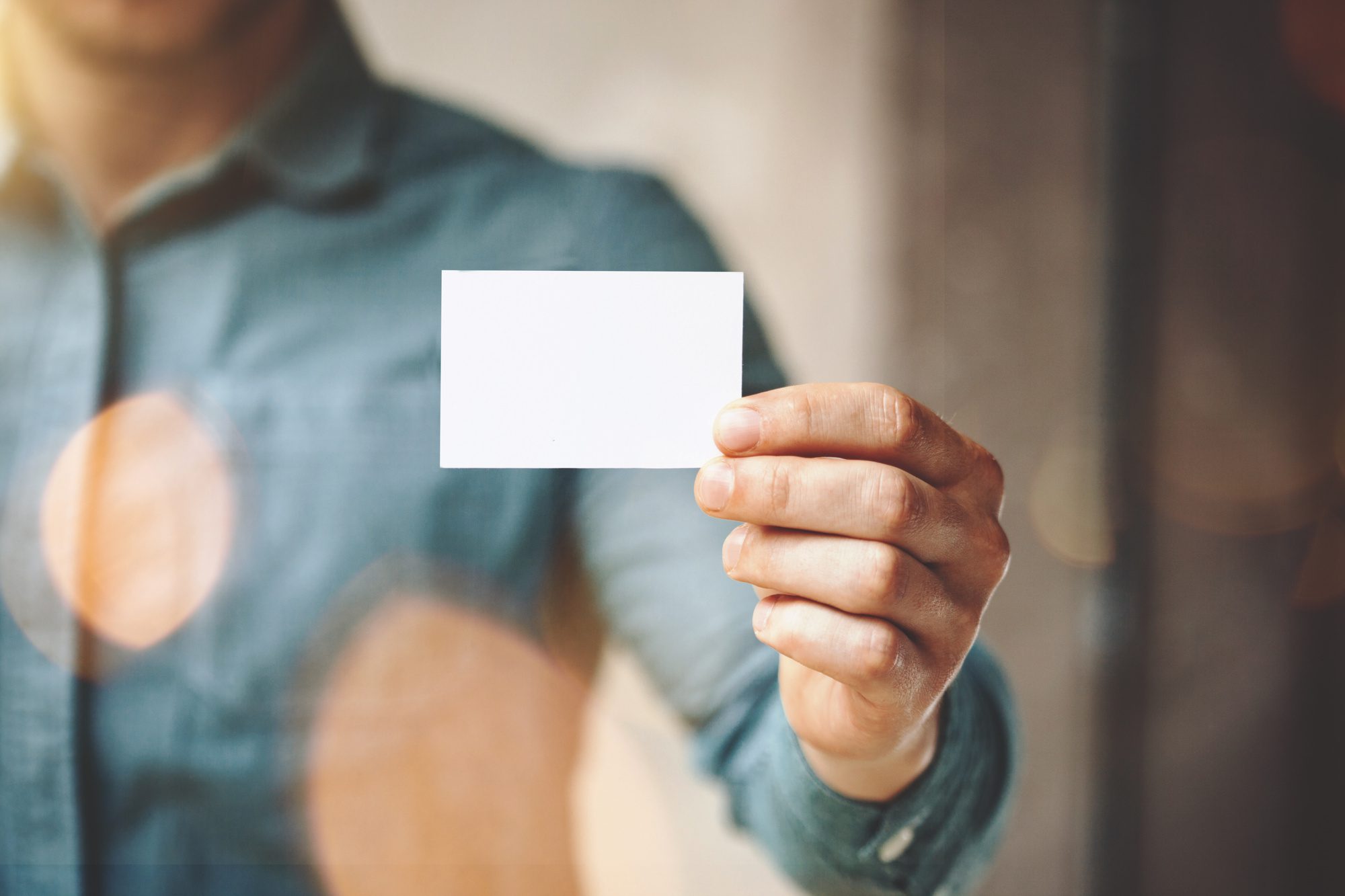 Man wearing blue jeans shirt and showing blank white business card Blurred background Horizontal mockup